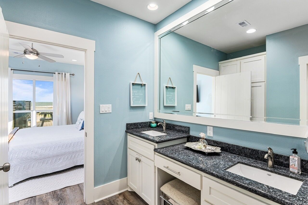 A bedroom with a bed, a bathroom with a large mirror, two sinks, and a ceiling fan.