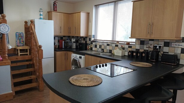 A compact kitchen with wooden cabinets, a built-in microwave, a washing machine, and a large window with blinds. - Roman Bank Flat With Spa Bath and Mermaids Teepee (Skegness)