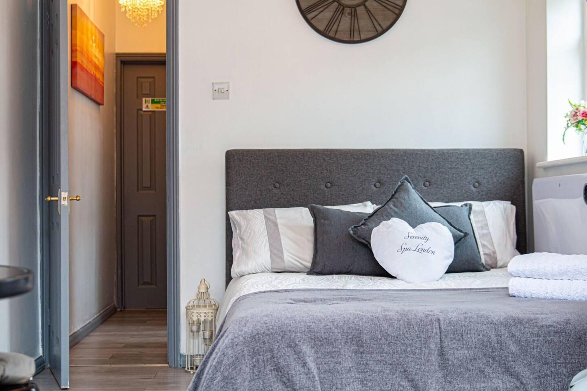A bedroom with a grey upholstered headboard, a bed with grey and white bedding, and a white heart-shaped pillow with text.