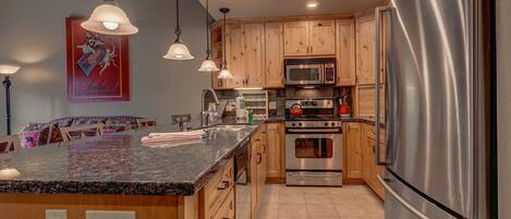 A kitchen with wooden cabinets, stainless steel appliances, granite countertops, pendant lighting, and a tiled floor.