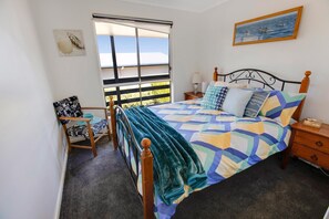 Een slaapkamer met een bed, een stoel, een nachtkastje, een raam en een schilderij aan de muur.