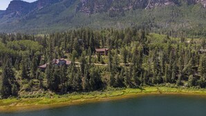 Una cabana a la vora d'un llac, envoltada per un bosc dens i muntanyes.