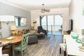 Un grand espace de vie comprenant une table à manger, des chaises, un canapé et un téléviseur. Un balcon offre une vue sur l’océan.