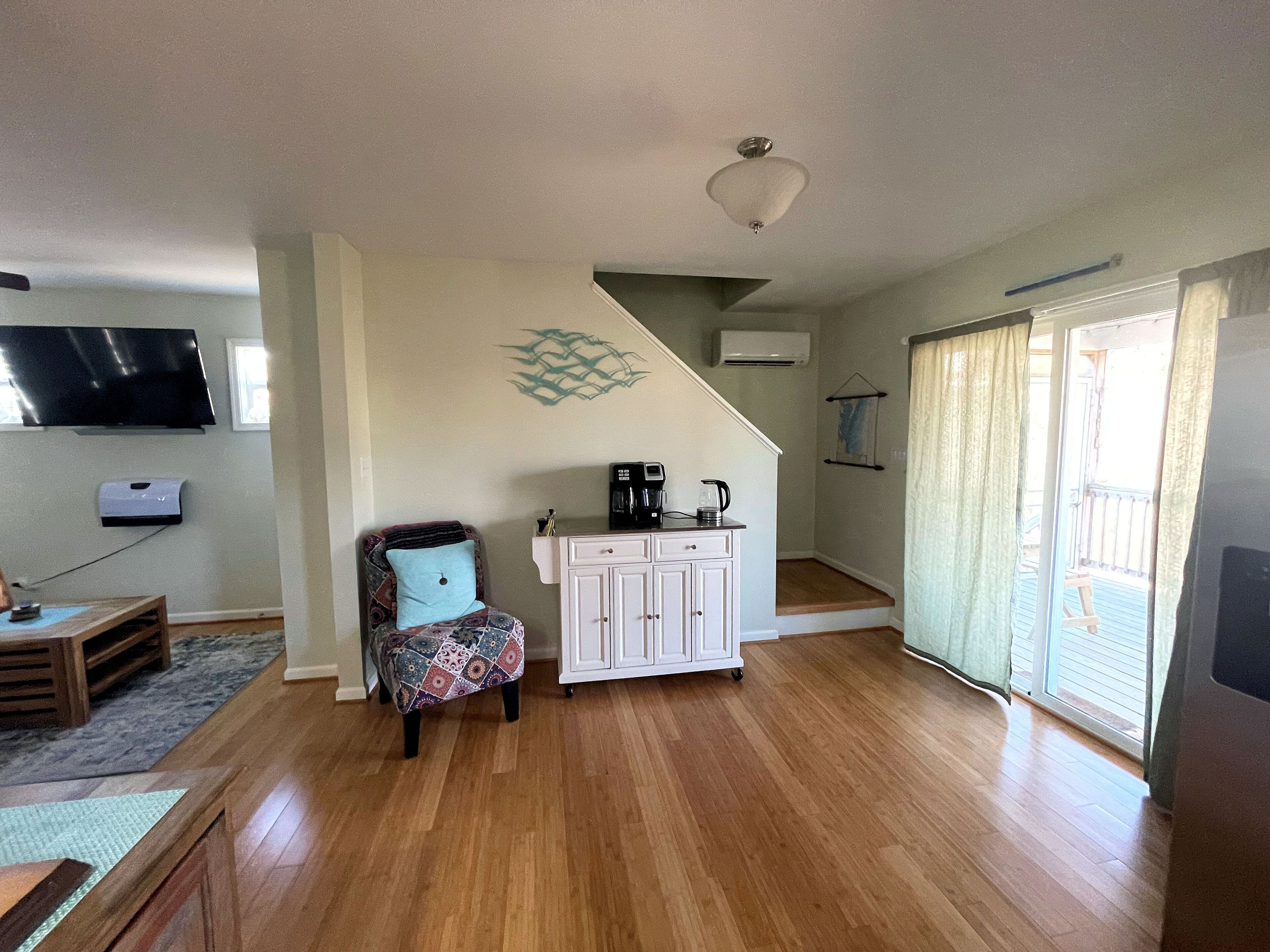 Una sala de estar con piso de madera, un sillón estampado, un gabinete blanco, una cafetera y un televisor montado en la pared.