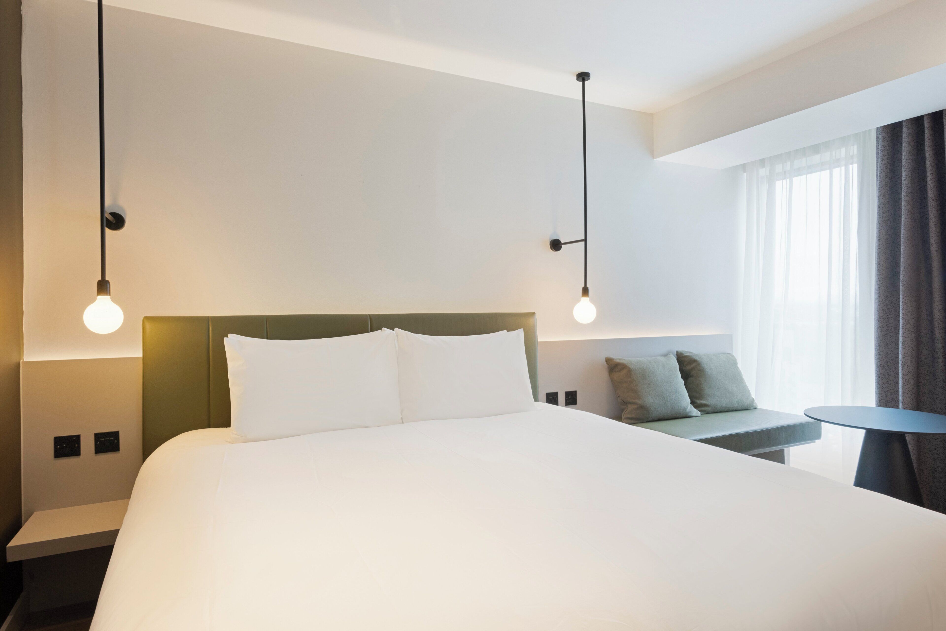 A hotel room with a large bed, a bedside table, a small round table, and a window with curtains.