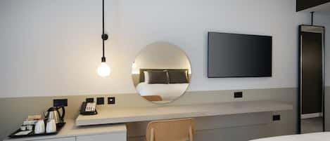 A modern hotel room with a bed, a desk with a chair, a round mirror, a flat-screen TV, and a coffee station.