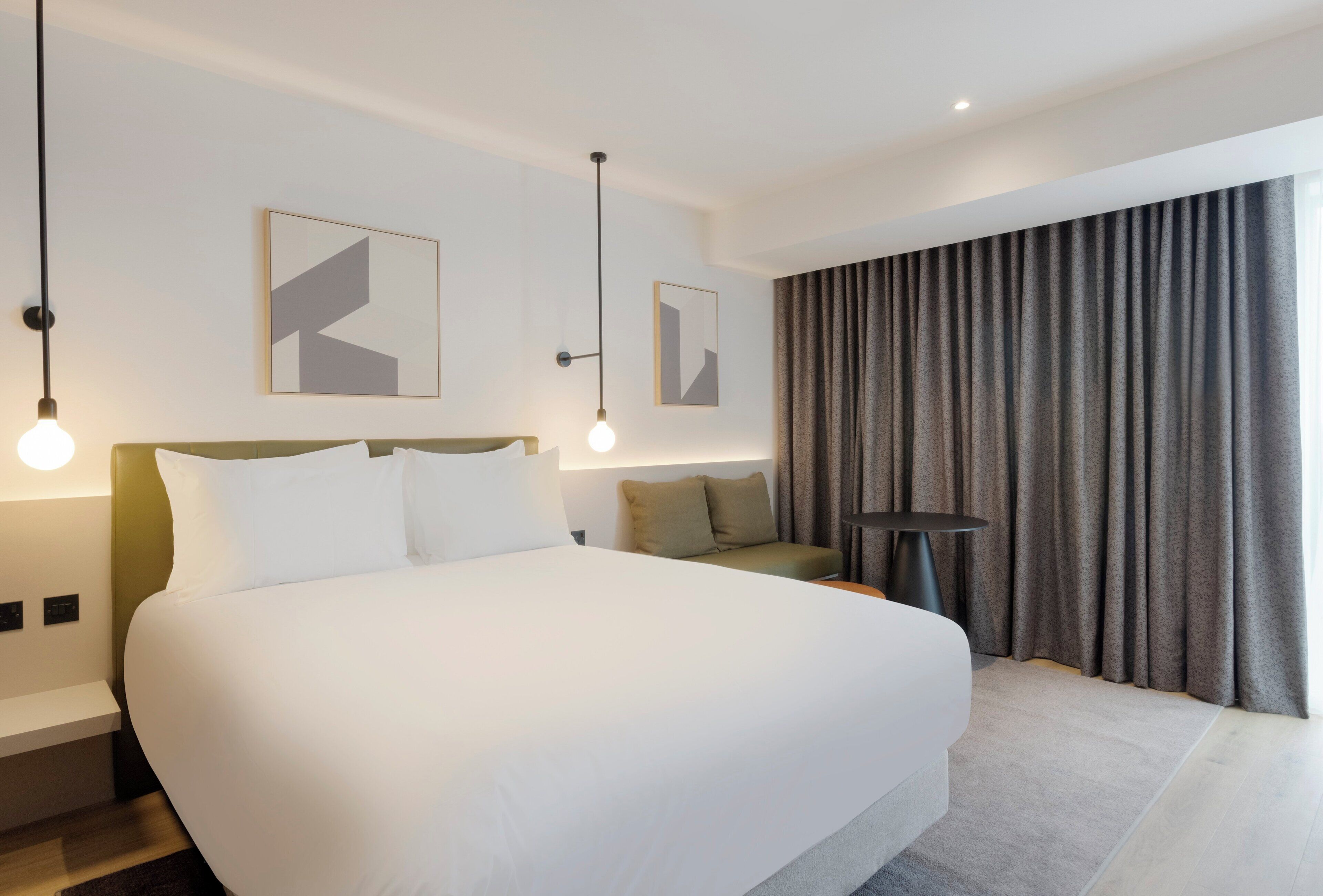 A modern hotel room with a large bed, a sofa, a small table, and two wall-mounted light fixtures.