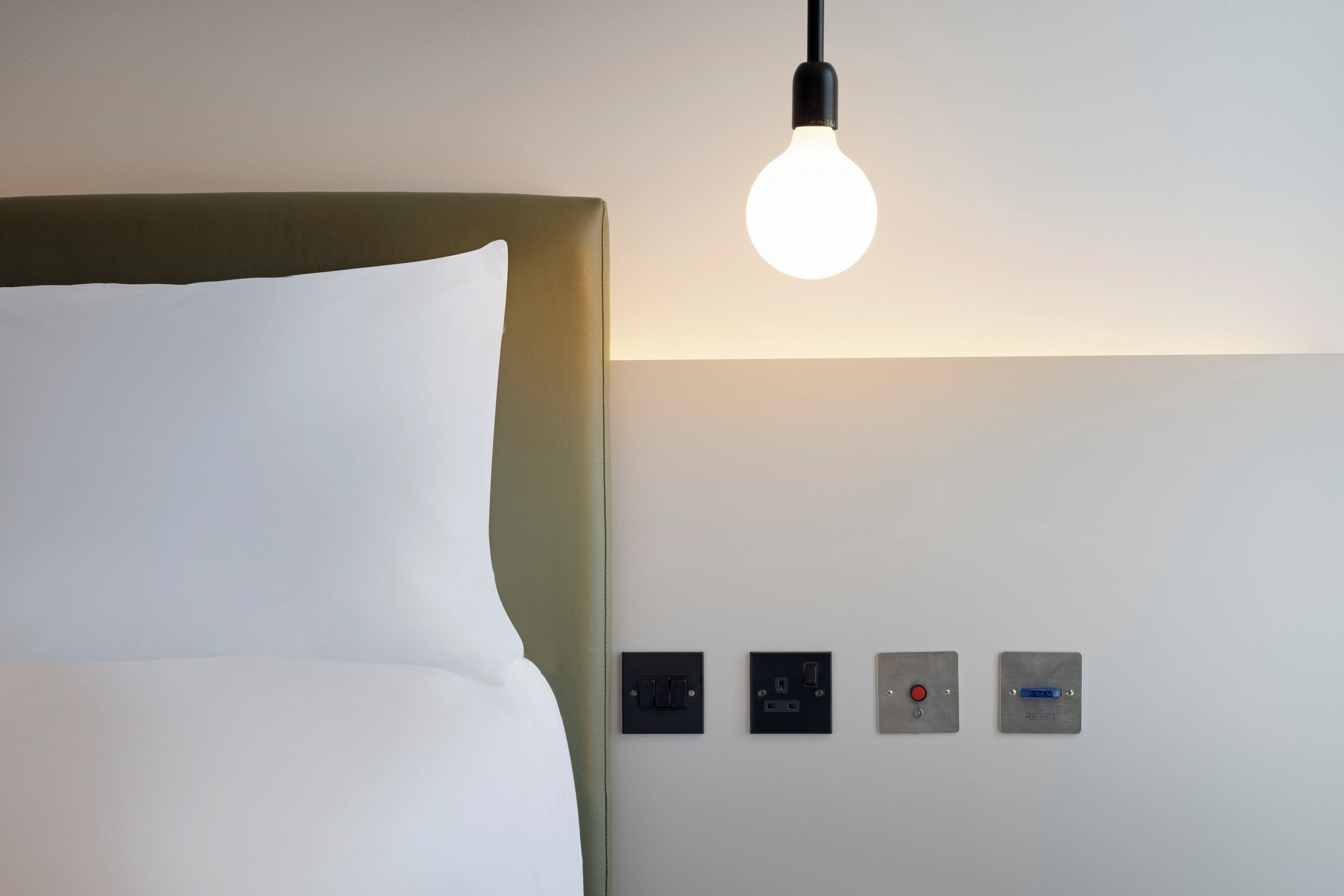 Ein Hotelzimmer mit einem Bett, einer Wandleuchte und einer Bedienkonsole mit Knöpfen und einem roten Knopf.