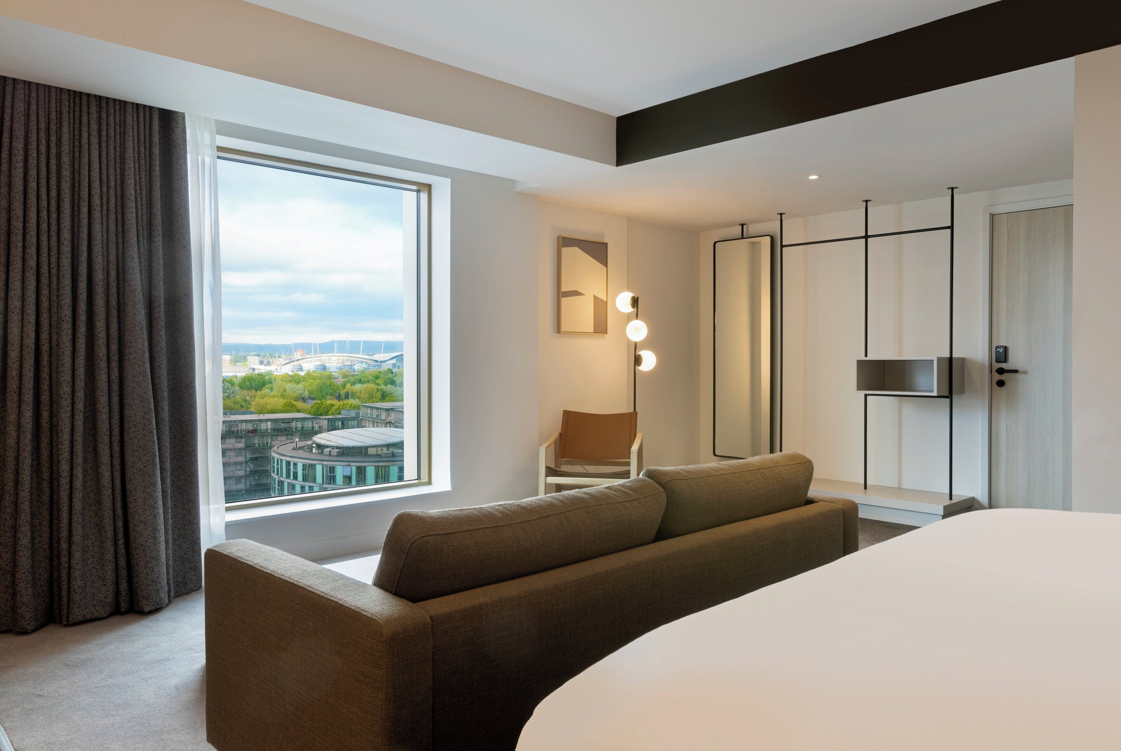 A modern hotel room with a large bed, a seating area with a sofa, a chair, and a desk. There is a large window with a view of a cityscape and a balcony.
