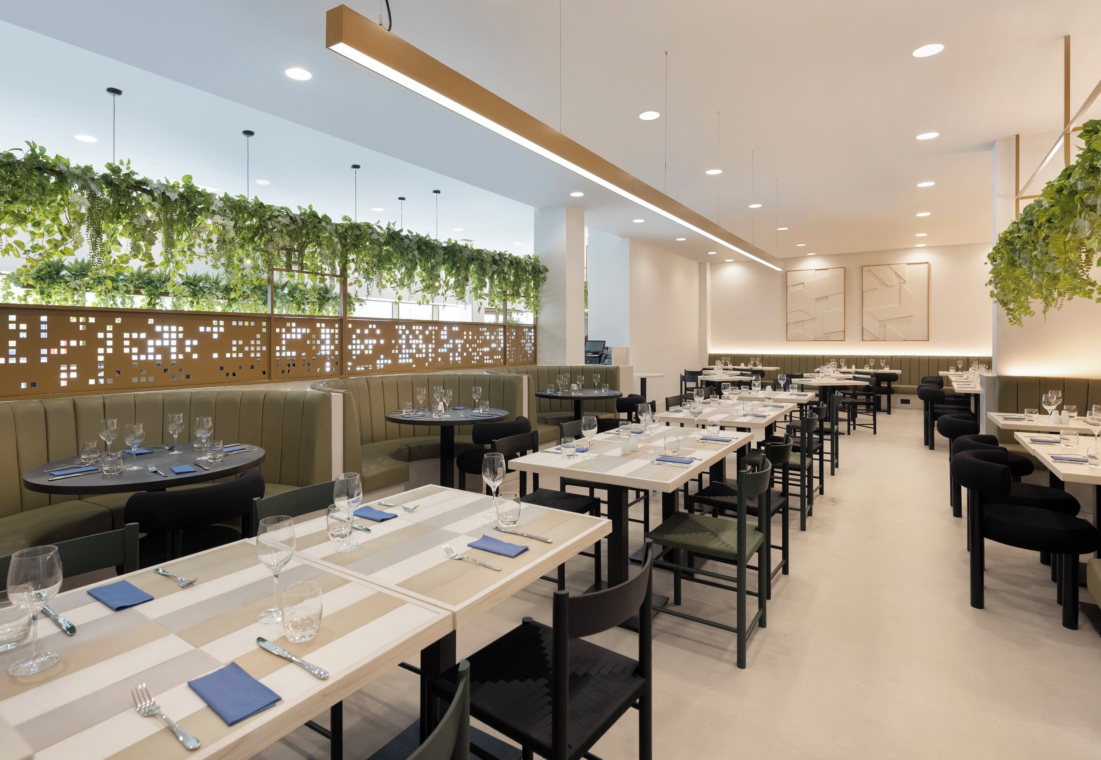 Ein modernes Restaurant mit Sitzgelegenheiten in Form von Boxsitzbänken und Tischen, einer grünen Pflanzenwand und abgehängten Deckenleuchten.