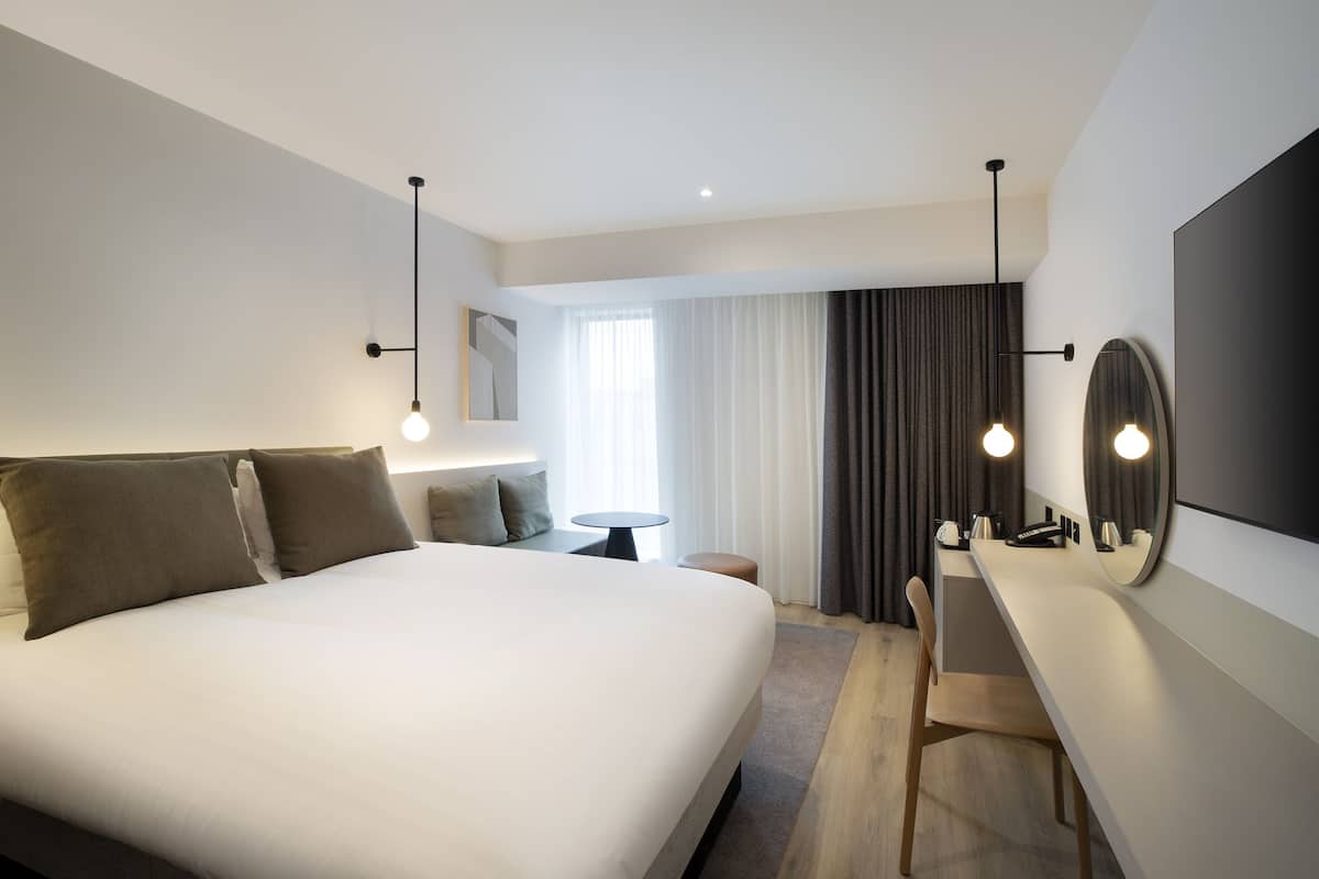 A modern hotel room with a large bed, bedside table, mirror, and a view of the outdoors.