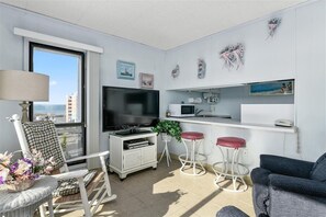 Uma sala de estar moderna com televisão de tela plana, uma área de jantar com mesa branca e uma cozinha com micro-ondas.