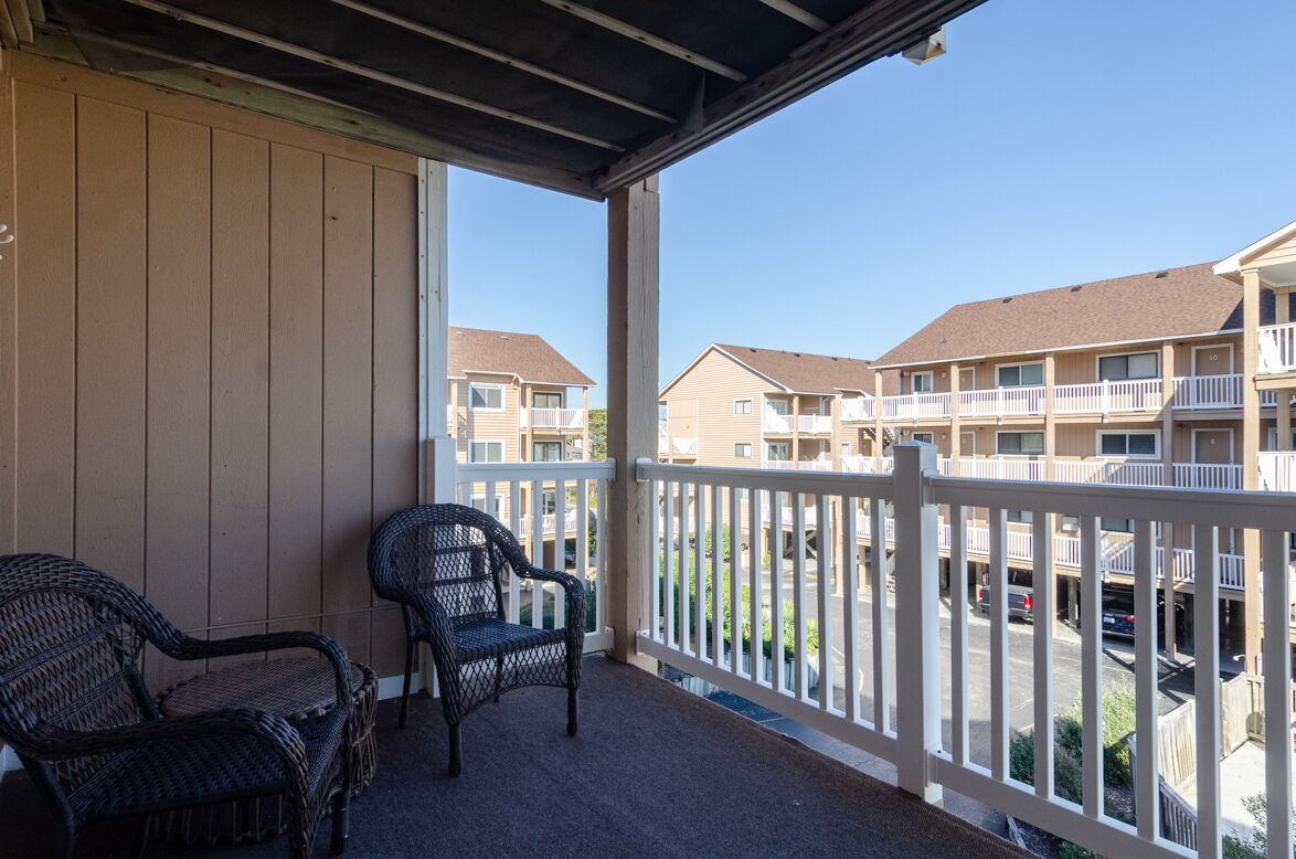 A balcony with wicker chairs, a view of multi-story buildings, and a parking area.