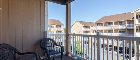 A balcony with wicker chairs, a view of multi-story buildings, and a parking area.