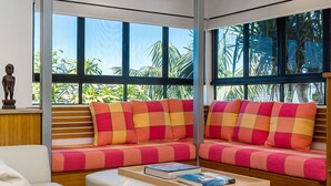 A modern room with large windows, a sofa with colorful cushions, a small table with books, and a wooden bench. - Pet-friendly 4BR coastal home with water views, timber deck and linen included (Evans Head)