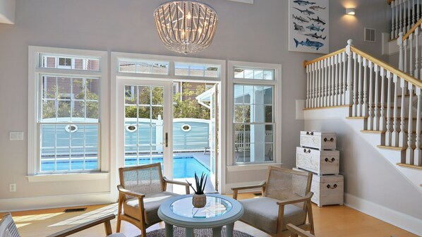 Relax in a spacious room with a high ceiling, featuring a large window with a view of the outdoor pool and a comfortable seating area with wicker chairs.
