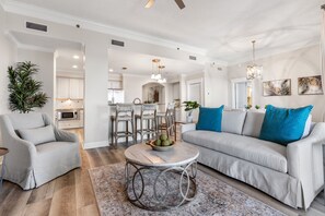 Uma sala de estar moderna com um sofá cinzento, uma mesa de centro redonda e um espaço de refeições com cadeiras, com uma cozinha ao fundo.