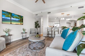 Uma sala de estar moderna com um quadro grande de uma praia, uma mesa de centro redonda e um sofá com almofadas azuis.