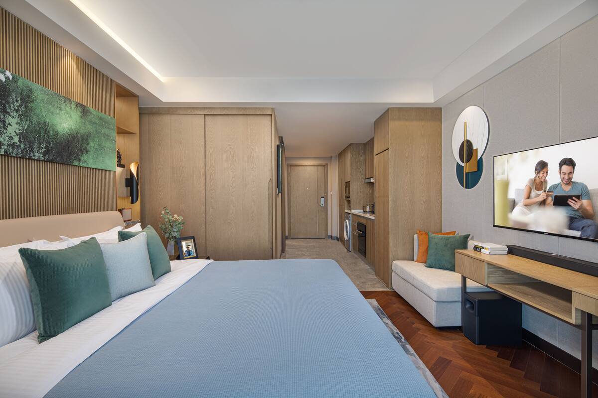 Une chambre d’hôtel moderne équipée d’un grand lit, d’une télévision, d’un bureau et d’un canapé. Une porte donne accès à une salle de bain et à une kitchenette.