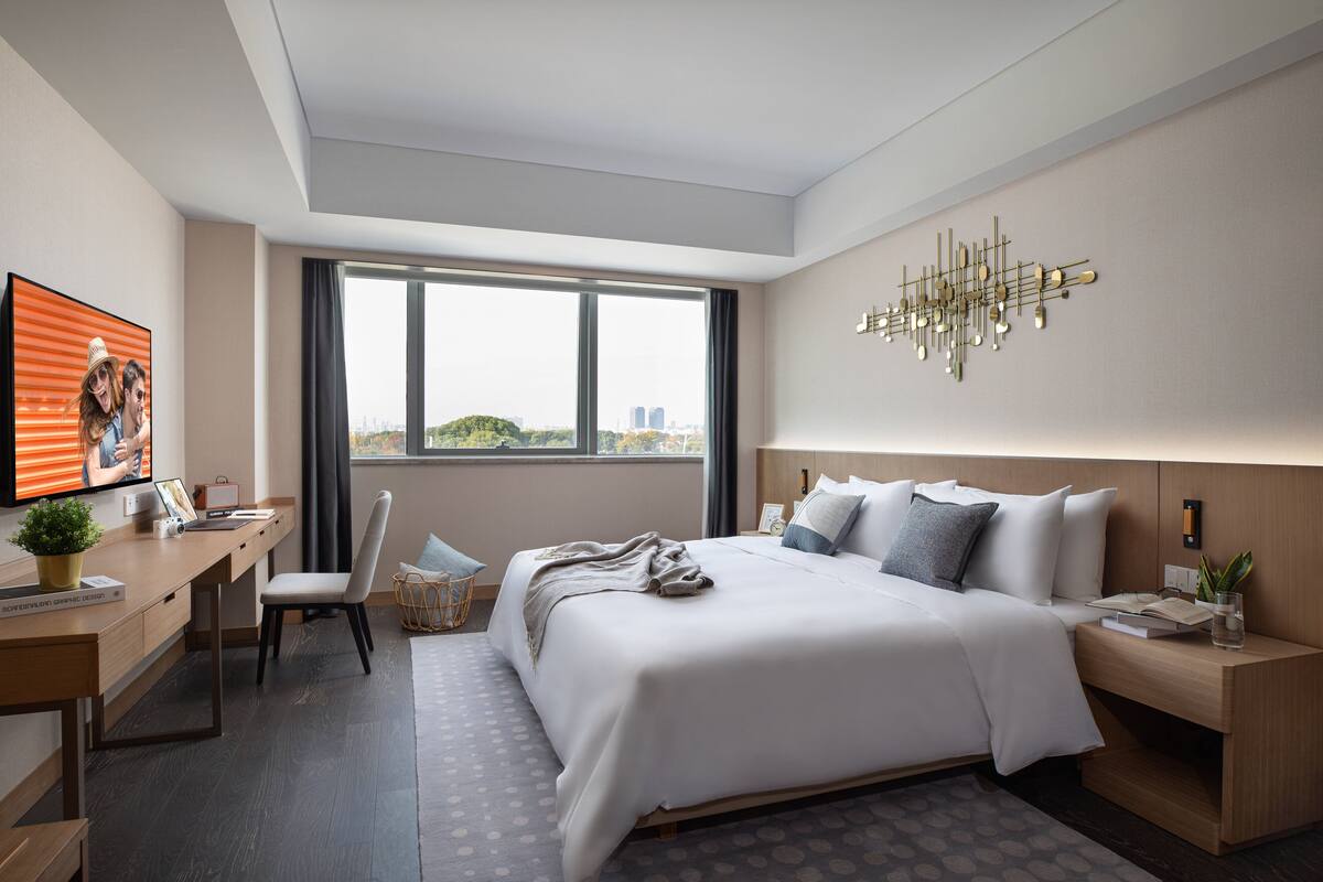 A modern hotel room with a large bed, a desk with a computer, a chair, and a view of the city through large windows.
