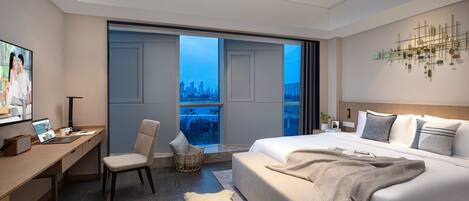 A modern hotel room with a large bed, a desk with a laptop, a chair, and a view of the cityscape through large windows.