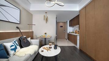 A modern living room with a sofa, coffee table, and a guitar. There's a dining area with a table set for two and a kitchen with wooden cabinets.