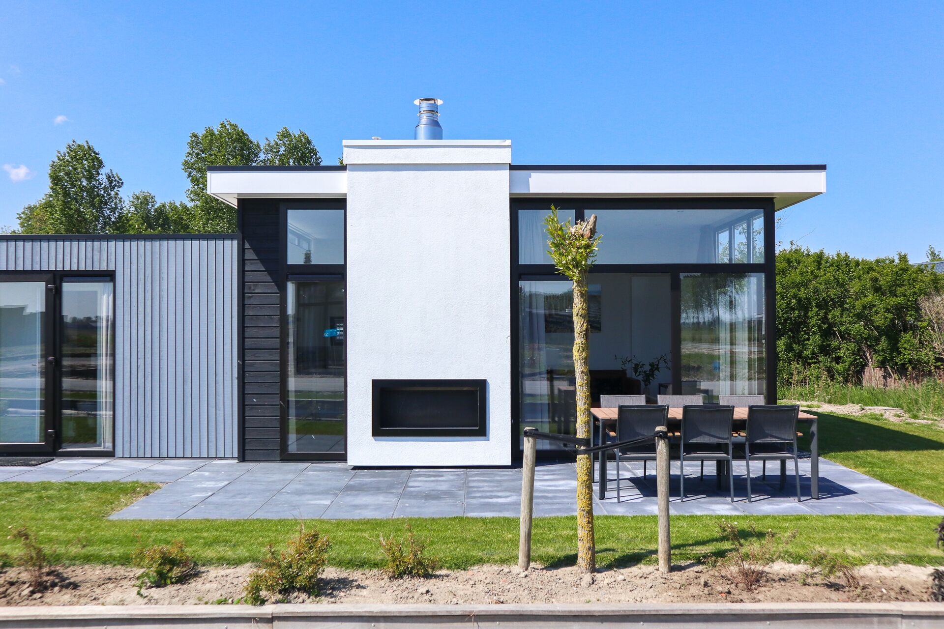 A modern two-story house with a large glass facade, a central chimney, and an outdoor seating area with chairs and a table.