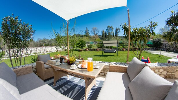 An outdoor patio with wicker furniture, a table with a drink, and a view of a garden and pool. - Giannakis Villa Corfu (Corfu)