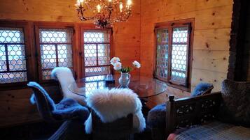 A cozy wooden cabin interior with a round glass table, wicker chairs, a chandelier, and large windows with wooden frames.