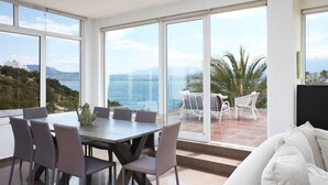 A modern dining area with a table and chairs, large windows with a view of the sea and mountains, and a balcony with outdoor furniture. - Longobardi Sea Front Villa Chania Greece (Chania)