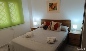 A hotel room with a bed, two bedside tables, a window with blinds, a painting on the wall, and a door. - Apartment Huerta del Obispo (Cádiz)