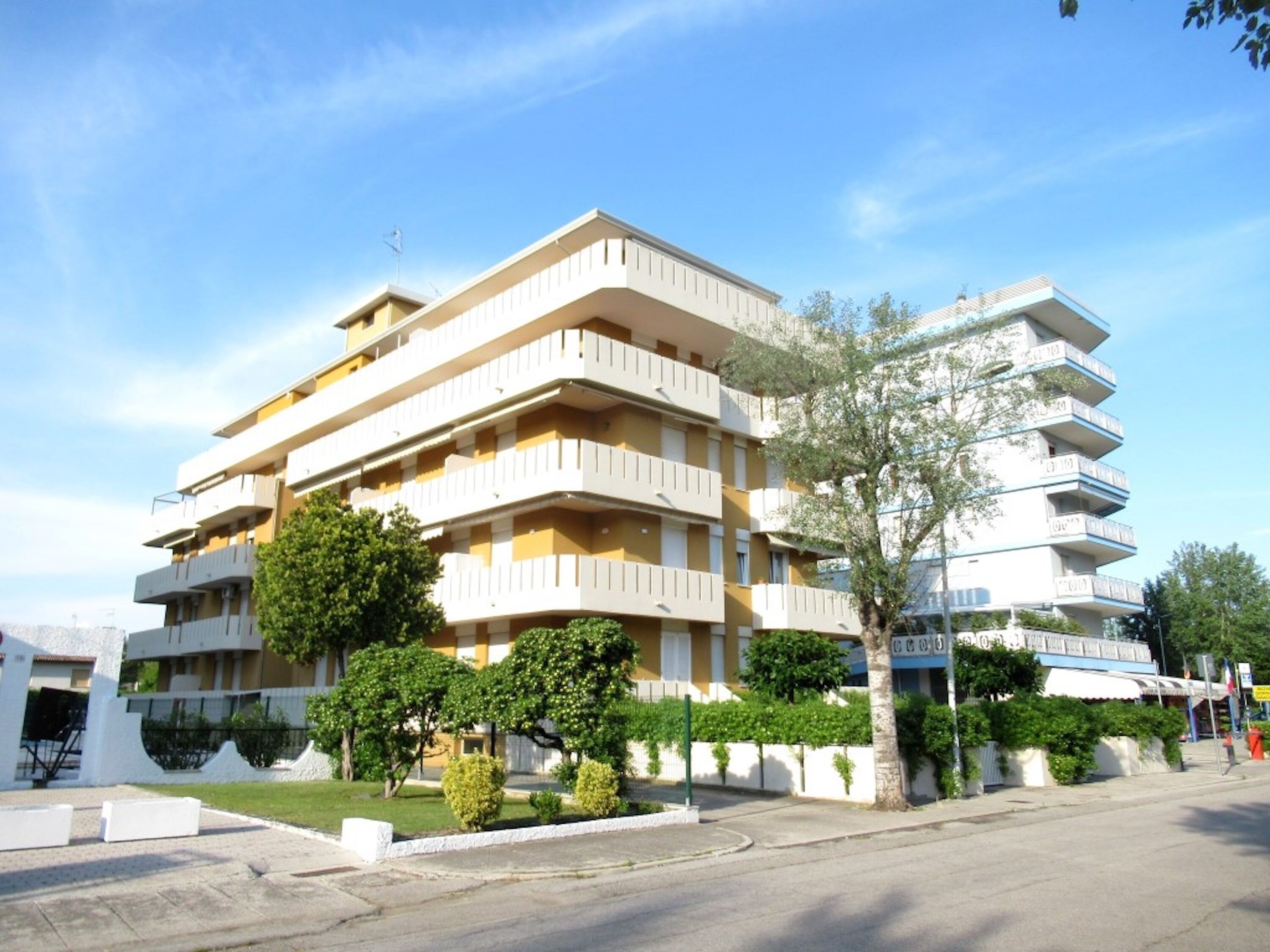 A multi-story residential building with balconies, surrounded by greenery and a clear sky.