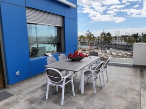 Eine Terrasse mit Tisch und Stühlen, Blick auf den Strand, und eine blaue Gebäudefassade.