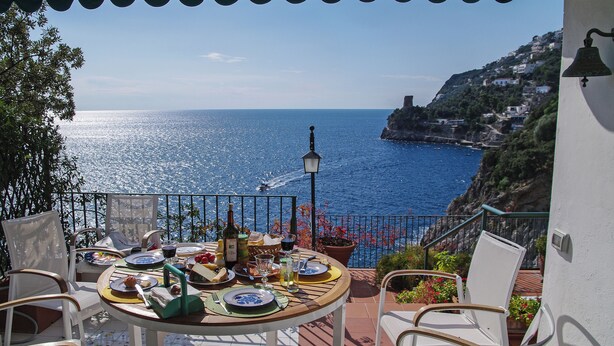 Eine Terrasse mit Esstisch, die auf eine Küstenlandschaft mit Häusern an einem Hügel blickt.
