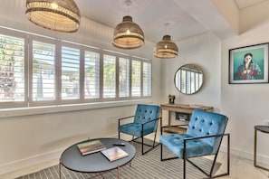 Una habitación con ventanas amplias, lámparas colgantes de mimbre, un espejo redondo y sillones tapizados en azul.