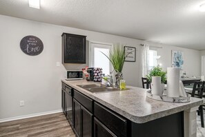A modern kitchen with dark cabinetry, a granite countertop, and a central island. There is a microwave, a coffee maker, and a vase with plants on the island. - BMT GRADUATION 🔥 5MN TO LACKLAND AFB 🔥 DISABLED VETERAN OWNED (San Antonio)