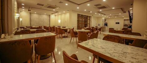 A spacious dining area with marble-topped tables and brown chairs, featuring recessed ceiling lighting and a kitchen area in the background.