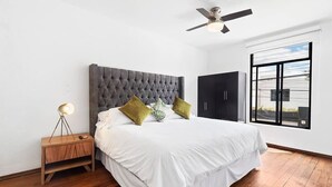 Un dormitorio con cabecera capitonada, cama con ropa de cama blanca, dos almohadas verdes, mesita de noche de madera con una lámpara y un armario negro.