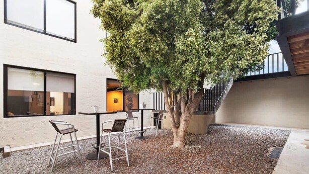 Un área de estar al aire libre con una mesa, sillas, un árbol y un edificio con ventanas grandes.