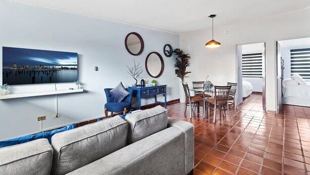 Ein modernes Wohnzimmer mit einer Couch, einem Esstisch mit Stühlen, einem an der Wand montierten Fernseher und einem großen Fenster.