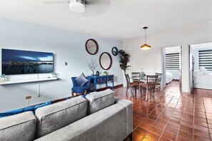 A modern living room with a sofa, a dining table with chairs, a TV mounted on the wall, and a large window.