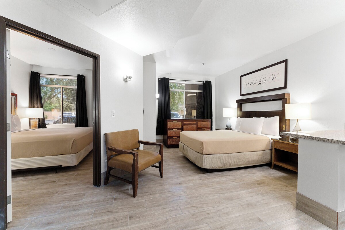 A hotel room with two beds, a chair, a dresser, and a desk with a lamp.