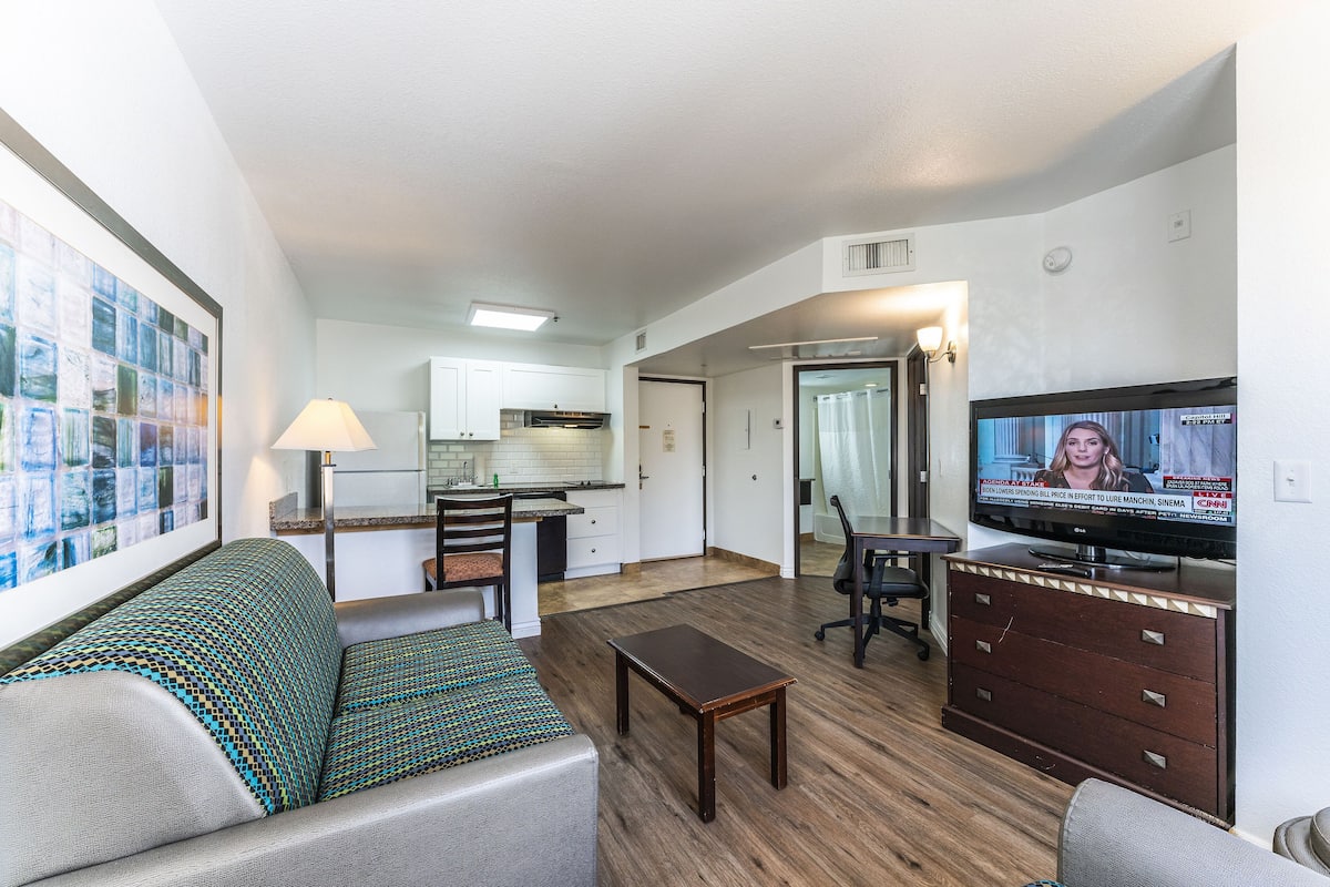 A living room with a sofa, a coffee table, a TV, and a kitchen in the background.
