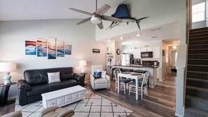 A modern living room with a black leather sofa, a white coffee table, and a dining area with a white table and chairs. There is a ceiling fan, a kitchen in the background, and a staircase to the right. - "Coastal Vibes" One Block from Beach-Unit 1011-Island Dunes Condominiums (Port Aransas)