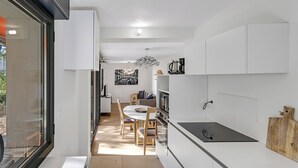 Uma cozinha moderna com armários brancos, uma área de refeições com mesa e cadeiras redondas, e vista para o exterior por uma grande janela.