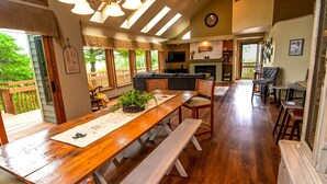A spacious living area with a wooden dining table, benches, and a sofa. A fireplace and a television are visible in the background. - Wow Sunsets/Hot Tub/Fab Game Room/Fire Pit! Close to Owners' Club, Golf and Spa! (Galena)