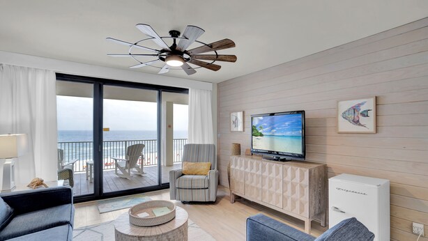 Una sala de estar moderna con un televisor grande, un ventilador de techo, un sofá, una silla y vista al océano a través de una puerta corrediza de vidrio.