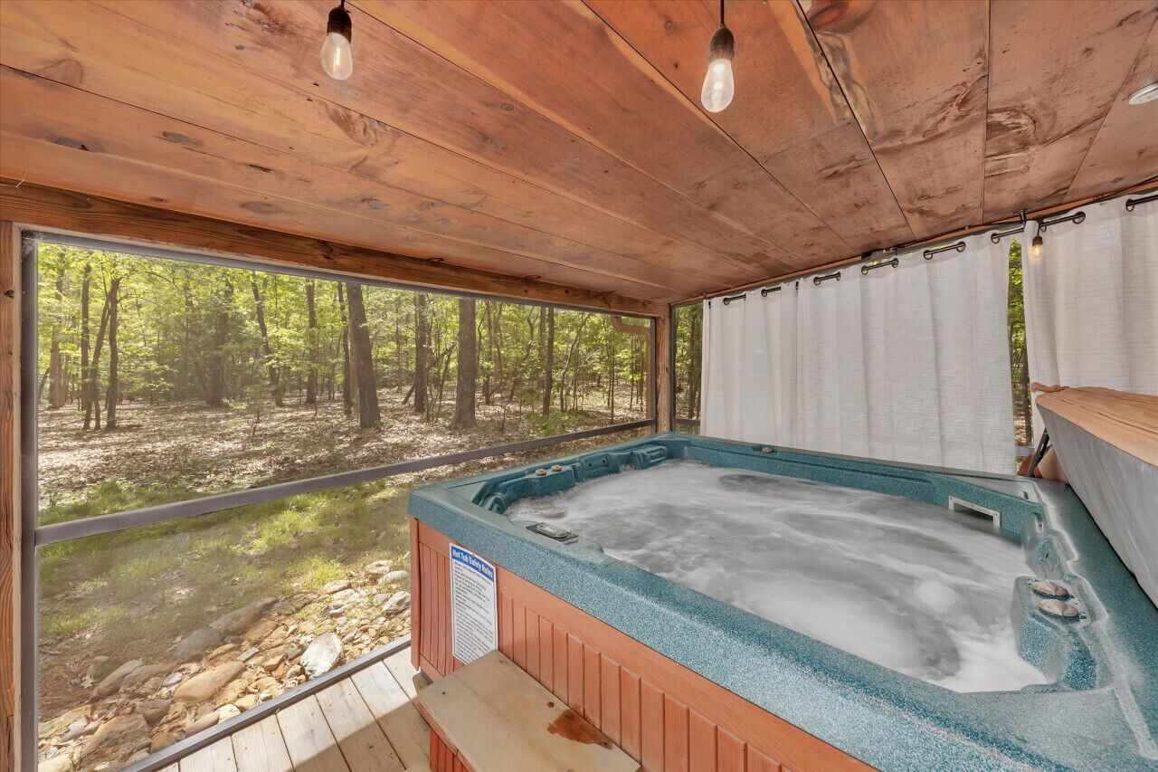 A wooden cabin with a hot tub, large windows, and a view of a forest.