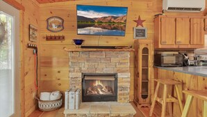 Un interior acogedor de cabaña con chimenea de piedra, paredes de madera, televisor montado y una zona de cocina con armarios y taburetes de madera.