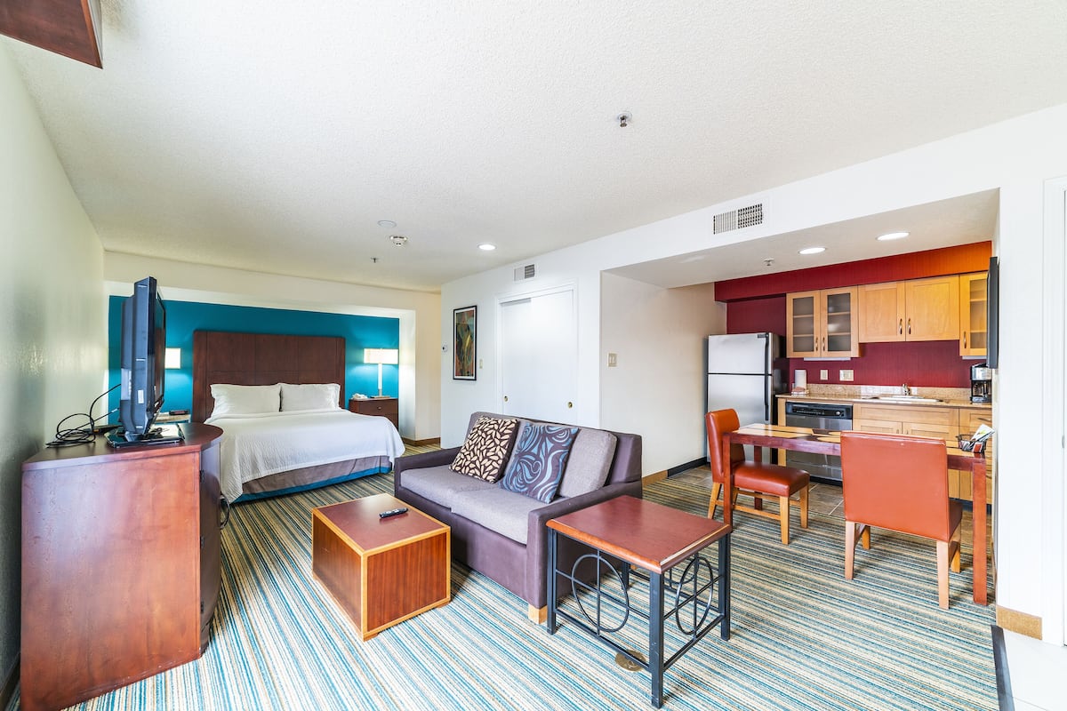 Quarto de hotel com cama, sofá, mesa de centro, televisão e uma cozinha compacta com geladeira, fogão e armários.
